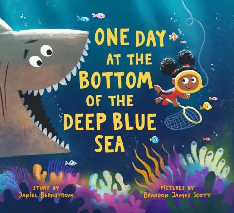 Photo of Daniel B's book called "One Day at the Bottom of the Deep Blue Sea." There is a shark smiling, preparing to chomp the yellow words. A girl is swimming on the other side of the text and smiling as well. 